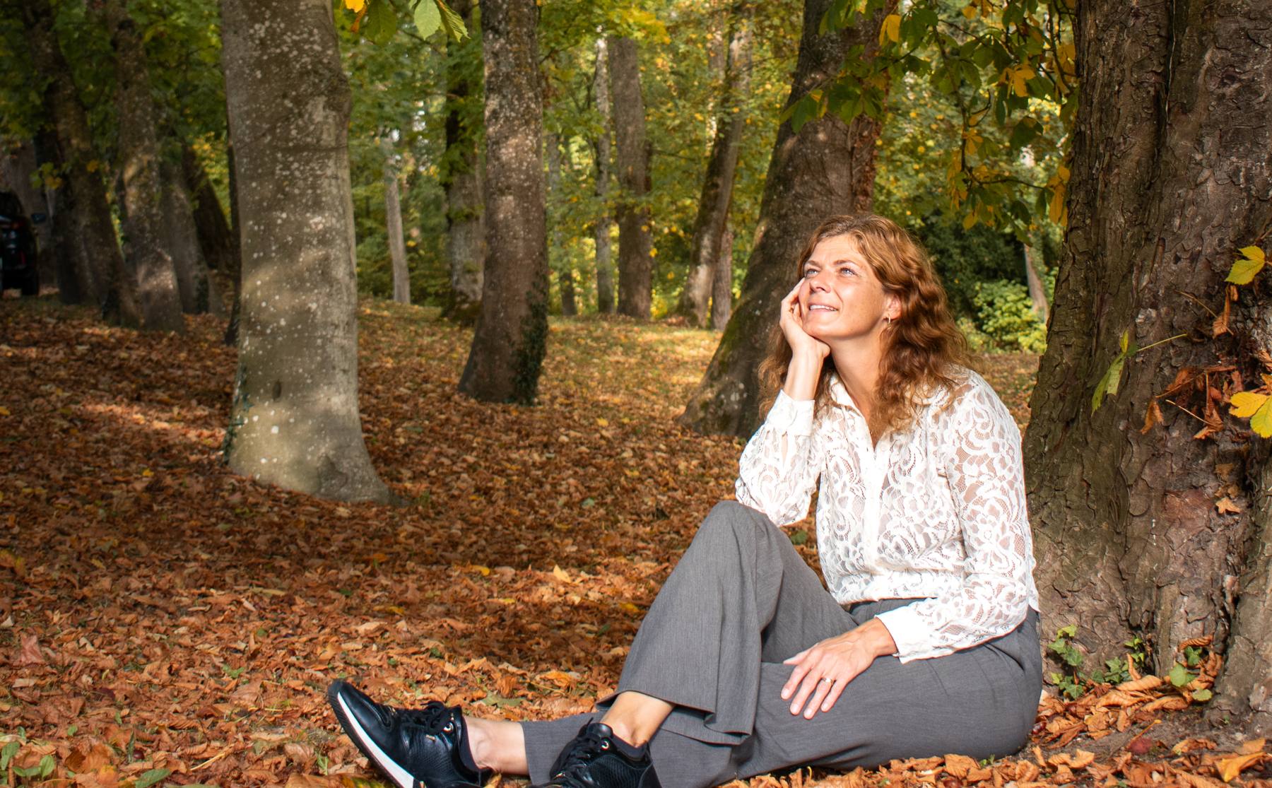 Jantine zit tegen een boom tussen de herfstbladeren en kijkt glimlachend omhoog